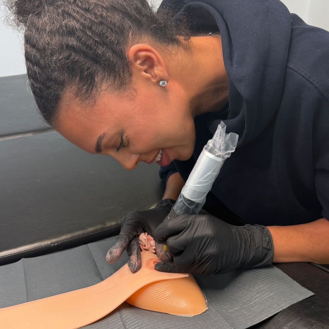 A paramedical tattoo artist in training practices 3D areola tattooing on synthetic skin, focusing on realistic nipple and areola restoration techniques. The artist wears gloves and uses professional tattoo equipment, learning advanced shading, scar camouflage methods, and paramedical tattoo skills that support women after mastectomy, breast reconstruction, or breast cancer recovery.