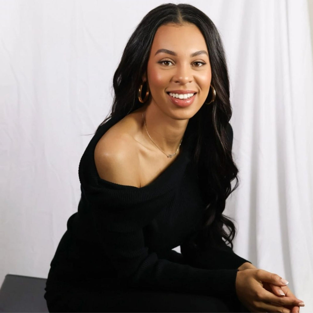 A smiling Alyssa with long dark hair wearing a black off-the-shoulder long-sleeve top, seated against a white draped studio background.